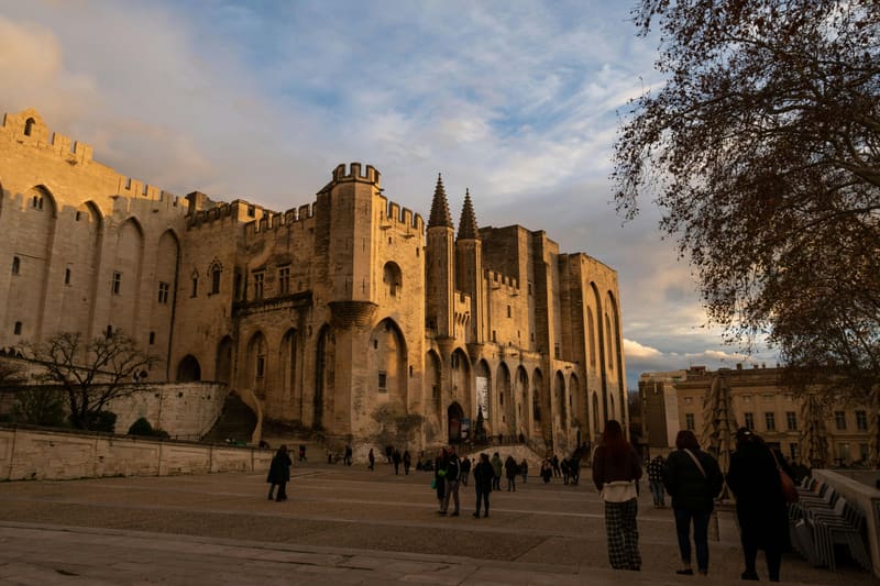 The Palais des Papes (Papal Palace) in Avignon at golden hour — visitors crossing the Place du Palais beneath the limestone walls and twin spires of the largest Gothic palace ever built in Europe. UNESCO World Heritage Site since 1995, seat of the Avignon Papacy 1309-1377.
